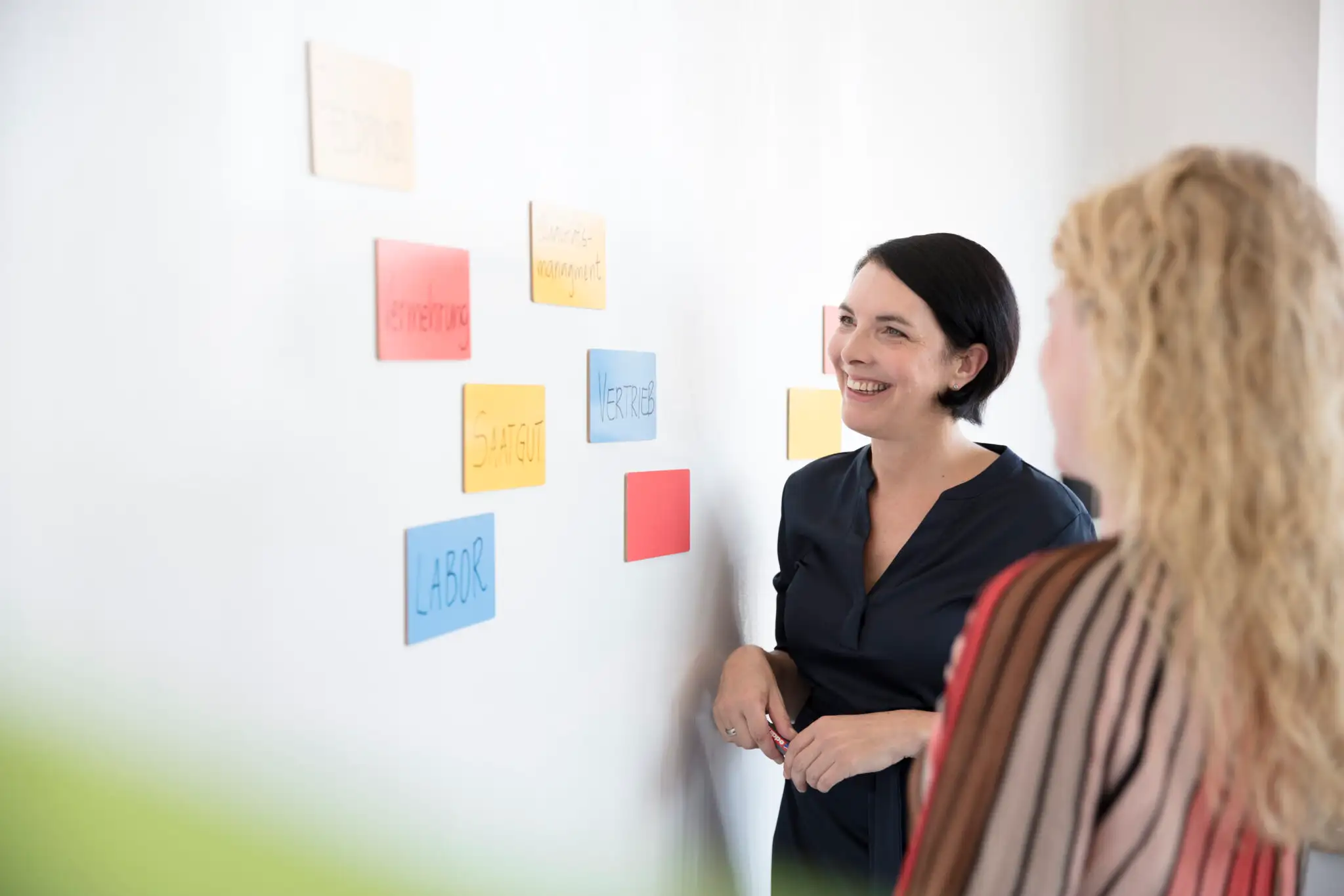 Zwei Frauen stehen vor einer Pinwand auf der Post Its hängen